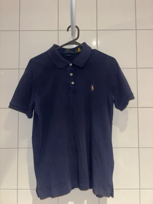 Marinblå pikétröja från Ralph Lauren - Klassisk marinblå pikétröja från Ralph Lauren med korta ärmar och krage. Tröjan har tre knappar framtill och den ikoniska broderade loggan på bröstet. Tillverkad i mjuk bomull som känns skön mot huden, inte strykt så lite strynklig!