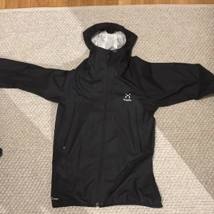 Svart vindjacka från Haglöfs, stl S - Svart regnjacka från Haglöfs i storlek S. Jackan har huva, dragkedja framtill och två sidofickor med dragkedja. Tillverkad i slitstark syntet med minimalistisk design och vitt Haglöfs-tryck på bröstet. Perfekt för blöta dagar och outdoor.