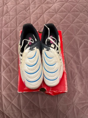 Puma vita fotbollsskor med blå detaljer - Säljer ett par vita fotbollsskor från Puma med blå och svarta detaljer samt rosa logga. Skorna har en klassisk snörning och mönstrad ovandel i syntetmaterial. Perfekta för dig som vill sticka ut på planen med färgstark design.