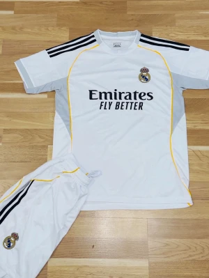 Real Madrid vit matchställ - Säljer ett vitt matchställ från Real Madrid med klubbens logga på både tröja och shorts. Tröjan har svarta och gula detaljer samt texten 'Emirates Fly Better' på bröstet. Shorts med svarta ränder och logga. Perfekt för fotboll eller träning.