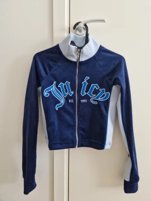Juicy Couture navy blue ziptröja XS - Mörkblå ziptröja från Juicy Couture med ljusblå detaljer och stor text 'Juicy' framtill. Tröjan har hög krage, långa ärmar och är tillverkad i mjukt velourmaterial. Perfekt för en sportig och trendig look. Limited edition i begränsat lager, storlek XS.