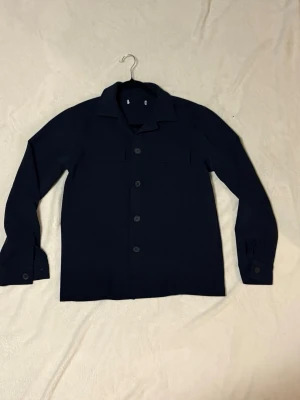 Mörkblå Overshirt från H&M - Säljer en stilren mörkblå overshirt från H&M med klassisk krage och knappar framtill. Skjortan har långa ärmar med knapp vid manschetten och är gjord i ett mjukt material som passar perfekt till olika outfits. Enkel och snygg design. Det är size M men hade även kunnat passa S fint med. 