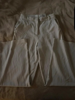 Vida beige kostymbyxa med pressveck - Stilrena kostymbyxor i beige med vida ben och pressveck. Byxorna har hällor för skärp, diskreta fickor bak och stängs med dragkedja och hake. Perfekta för en clean och modern look. Materialet känns mjukt och följsamt.