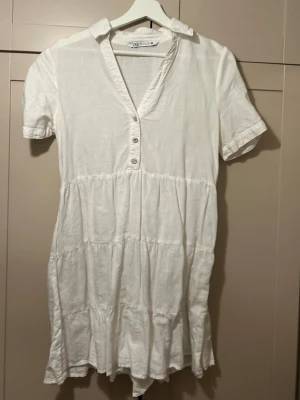 Vit kortärmad klänning, Zara - En luftig vit klänning från Zara med korta ärmar, krage och knappar framtill. Klänningen har en v-ringning.  Perfekt för sommarens varma dagar och enkel att styla.