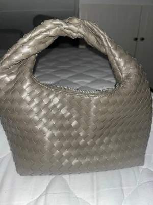 Beige flätad handväska med dragkedja - Trendig beige handväska i flätad design med kort, vadderat handtag och dragkedja upptill. Väskan har en mjuk, rundad form och är tillverkad i konstläder som ger en lyxig känsla. Perfekt accessoar för att lyfta din outfit. Köpte den för 395kr