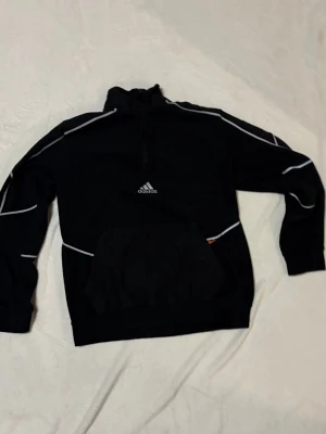 Svart hoodie från Adidas med reflex detaljer - Snygg svart hoodie från Adidas med reflex linjer längs ärmar och kropp. Halv dragkedja framtill, stor magficka och klassisk Adidas-logga på bröstet. Perfekt för en sportig och avslappnad stil. Tillverkad i mjukt material för extra komfort. Vid nacken finns lite böjningar som inte märks lika lätt. PRIS KAN DISKUTERAS 