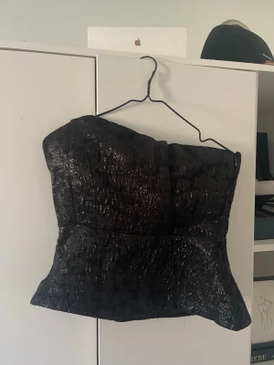 Svart glittrig korsett-topp - Säljer en svart korsett-topp med glittrig finish. Toppen är axelbandslös och har en strukturerad passform som framhäver midjan. Lite korsett vibe
