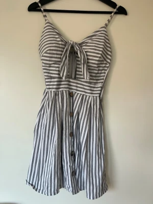Randig kort klänning  - Snygg kort klänning med smala axelband och knyt framtill. Klänningen har grå och vita ränder, knappar längs hela framsidan. Vadderad vid brösten.  Köpt utomlands så står ingen storlek men är som en S/M, använt fint skick! 