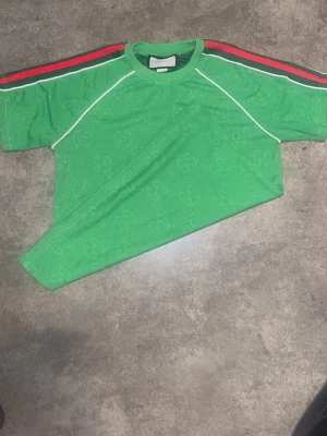 Grön Gucci t-shirt med ränder (äkta) - Snygg grön t-shirt från Gucci med diskret GG-mönster i tyget. T-shirten har korta ärmar och röda samt svarta ränder längs axlarna, samt vita kontrastsömmar. Perfekt för dig som vill sticka ut med en exklusiv streetstyle-look. Storlek S passar M, 100% äkta, super skick. 