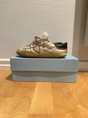 Vita sneakers från Philippe model - Snygga vita sneakers från Philippe modell med beige sula och bruna detaljer. Skorna har lågt skaft, klassisk snörning och svart hälparti. Tillverkade i läder med en cool vintage-look och unika sömmar på sidorna. Skorna är riktigt slitna och Box kommer inte med med om man vill ha med lådan så får man betala extra. Storlek 35 men passar 36.