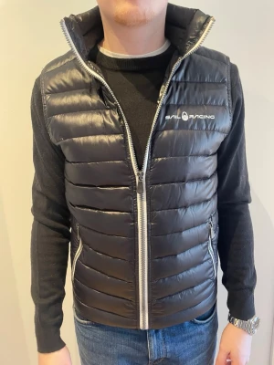 Svart dunväst från Sail Racing - Snygg svart dunväst från Sail Racing med hög krage och vit dragkedja. Västen har två fickor med dragkedja och diskret logga på bröstet. Quiltad design och lätt glansigt material ger en sportig look. Perfekt att slänga över en hoodie eller tröja.