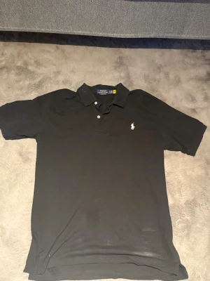 Svart pikétröja från Polo Ralph Lauren - Klassisk svart pikétröja från Polo Ralph Lauren med vit broderad logga på bröstet. Tröjan har krage, knappar framtill och korta ärmar. Perfekt för en stilren och avslappnad look.