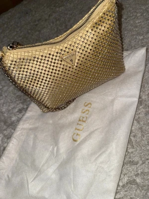 Guldig axelremsväska från Guess - Zalina shoulder bag från Guess i guld med glittrande stenar och metallkedja. I nyskick 