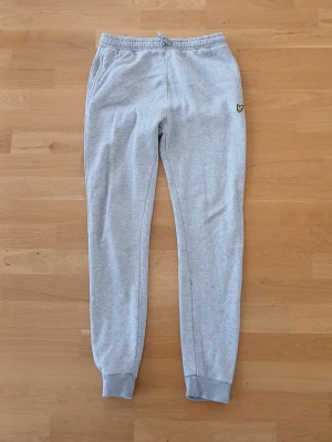 Grå mjukisbyxor från Lyle & Scott - Säljer ett par ljusgrå mjukisbyxor från Lyle & Scott med elastisk midja och muddar vid benslut. Byxorna har fickor på sidorna och en bakficka samt den klassiska Lyle & Scott-loggan broderad på framsidan. Perfekta för chill och avslappnade dagar. Står på byxorna att de är för 15/16 år men skulle rekomendera för 14/15 år beroende på längd. Längden på byxorna är 100 cm. Nästan oanvänd och ser ny ut.