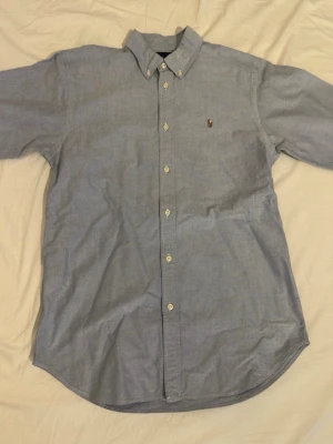 Blå skjorta från Ralph Lauren - Säljer en klassisk blå skjorta från Ralph Lauren med button-down krage och broderad logga på bröstet. Skjortan har långa ärmar och knappar framtill. Perfekt för dig som gillar stilrena plagg med ikonisk design.