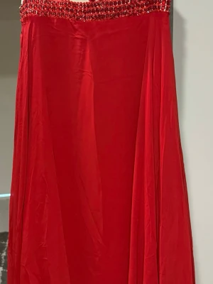 Röd långklänning med paljetter - Elegant röd långklänning med detaljerad paljettdekor upptill i rött på en ljus bas. Klänningen har en rund halsringning och ett flödande tyg som ger ett dramatiskt intryck. Perfekt för dig som vill sticka ut med en unik och glittrig look.