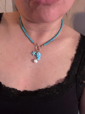 Turkost pärlhalsband med berlocker - Säljer ett trendigt halsband med små turkosa pärlor och guldfärgade detaljer. Halsbandet har tre berlocker: en blå evil eye, en stjärna och en vit pärla. Perfekt för dig som gillar färgglada accessoarer och vill sticka ut lite extra.