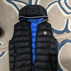 Svart dunväst med huva från Moncler - Jag har lappat för små hålen som knappt är synliga men det är en snygg svart dunväst från Moncler med blått foder och huva. Västen har två dragkedjefickor, quiltad design och klassisk Moncler-logga på bröstet. Perfekt för lager-på-lager och streetstyle. Stängs med dragkedja framtill. hål på framsidan men syns knappt 