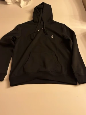 Svart hoodie från Polo Ralph Lauren - Snygg svart hoodie från Polo Ralph Lauren med klassisk huva och dragsnören. Har en känguruficka framtill och den ikoniska vita loggan broderad på bröstet. Perfekt för en chill och stilren look.