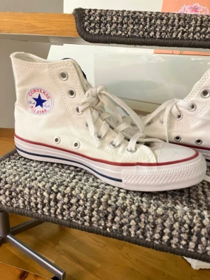Vita Converse All Star - Klassiska vita Converse Chuck Taylor All Star high tops med röd och blå rand på sulan. Skorna har canvasmaterial, vita skosnören och den ikoniska loggan på sidan. Perfekta för en avslappnad och cool streetstil.