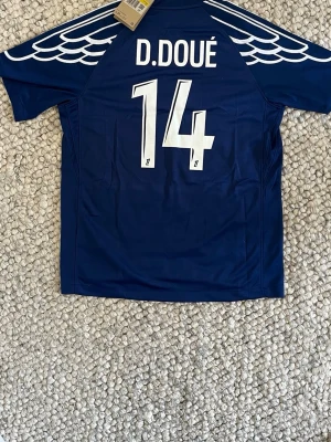 PSG x Jordan blå matchtröja - Mörkblå PSG matchtröja i samarbete med Jordan, med vita detaljer och vingmönster på axlarna. Tryck med D. Doué och nummer 14 på ryggen. Framsidan har PSG-logga, Jordan-logga och Qatar Airways sponsortryck. Priset kan diskuteras vid snabb affär🚚