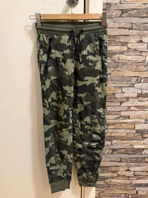 Camo mjukisbyxor med dragkedjor - Snygga mjukisbyxor i camouflage-mönster med svarta dragkedjor på fickorna och mudd vid benslut. Byxorna har snörning i midjan och en bakficka. Perfekta för en avslappnad och trendig stil.