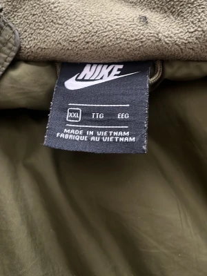 Olivgrön jacka från Nike XXL - Snygg olivgrön jacka från Nike i storlek XXL. Jackan är tillverkad i 100% polyester och har en sportig look med mjukt foder. Perfekt för dig som gillar streetwear och vill ha en stilren jacka med klassisk Nike-logga.