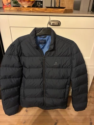 Svart pufferjacka från GANT - Snygg marinblå pufferjacka från GANT med diskret logga på bröstet. Jackan har en klassisk quiltad design, dragkedja framtill och blått innerfoder. Perfekt för kalla vinterdagar och håller dig varm med stil. Använd ett fåtal gånger och har storleken L.