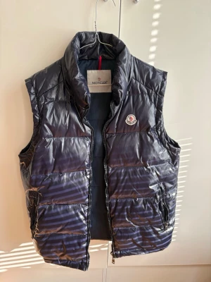 Mörkblå dunväst från Moncler - Snygg mörkblå dunväst från Moncler med hög krage och två dragkedjeförsedda fickor. Västen har ett glansigt yttertyg och klassisk Moncler logga på bröstet. Lite slitage i fickan och på dragkedjan, se bild. 