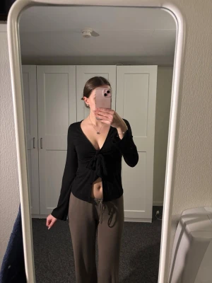 Svart omlottopp med knyt från Gina Tricot - Svart långärmad omlottopp från Gina Tricot med knyt framtill och djup v-ringning. Toppen är figurnära och har en croppad passform som visar magen. 