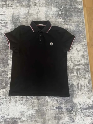 Svart pikétröja från Moncler - Svart pikétröja från Moncler med klassisk krage och knappslå framtill. 