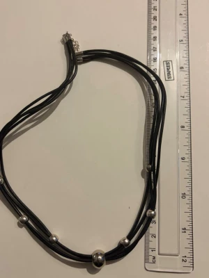 Svart halsband med silvriga pärlor - Stilrent halsband med flera svarta läderband och silvriga pärlor i olika storlekar. Halsbandet har ett metallspänne och en enkel, modern design som passar till många olika stilar.