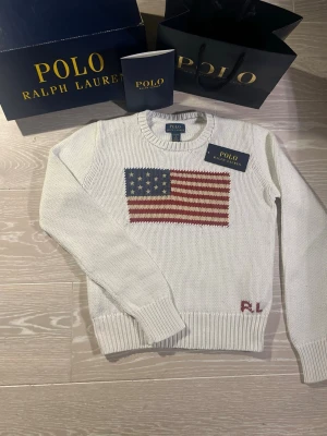 Vit stickad tröja Polo Ralph Lauren - Vit stickad tröja från Polo Ralph Lauren med amerikansk flagga i blått, rött och vitt på bröstet. Rund halsringning och ribbstickade muddar. RL-broderi i rött vid nederkanten. Klassisk och stilren design.Storlek L för 12-13 år 