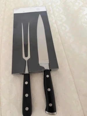 Trancherset kniv och gaffel - Snyggt trancherset med en kockkniv på 33 cm och en stekgaffel på 29 cm. Båda har svarta handtag med silverfärgade nitar och blad i rostfritt stål. Perfekt för att skära och servera kött på ett stilrent sätt.