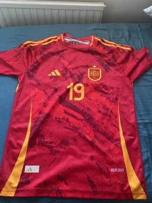 Spaniens matchtröja Adidas nr 19 - Röd och gul Spanien fotbollströja från Adidas med nummer 19 och namn Lamine Yamal på ryggen. Tröjan har gula detaljer, korta ärmar och är tillverkad i funktionsmaterialet HEAT.RDY. Perfekt för fotbollsträning eller att bära när du hejar på landslaget.
