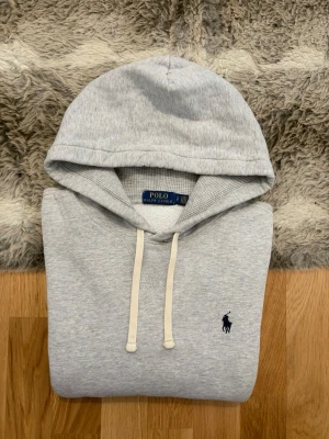 Ljusgrå Polo Ralph Lauren Hoodie - Storlek S - Hej! 👋 Säljer en klassisk ljusgrå Polo Ralph Lauren hoodie i storlek S. Den har den tidlösa marinblå pony-loggan broderad på bröstet 🐎 Tröjan är i gott skick, men jag vill vara helt ärlig med två små skönhetsfel: det finns ett litet märke/fläck på magfickan samt ett lagat hål vid midjan baktill (se närbilder). Det är inget som stör jättemycket när den bärs, men priset är satt därefter! ✨ 100% äkta med tvättlappar kvar. Perfekt som en skön och billig vardagshoodie! Fraktar snabbt! 🚛📦💬💯 