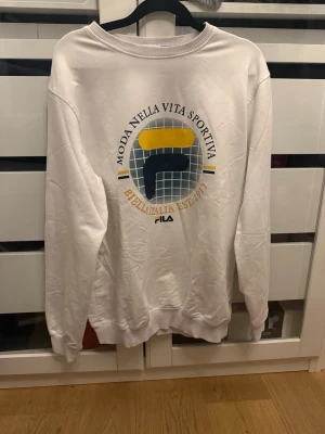 Vit sweatshirt från Fila med tryck - Vit sweatshirt från Fila med stort retroinspirerat tryck på ryggen i blått, gult och grått. Rund halsringning och ribbade muddar vid ärmslut och nederkant. Perfekt för en sportig och avslappnad stil. Passar M- XL
