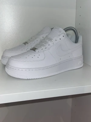 Nike Air Force 1 vita sneakers - Klassiska Nike Air Force 1 sneakers i helvitt läder med perforerad tå, vit sula och ikonisk Swoosh-logga på sidan. Skorna har lågt skaft och platt yttersula med cirkulärt mönster. Perfekta för en clean och stilren look.