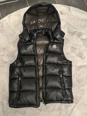 Svart dunväst från Moncler - Snygg svart dunväst från Moncler med avtagbar huva och två dragkedjefickor framtill. Västen har ett glansigt yttertyg och klassisk Moncler-logga på bröstet. Perfekt för lager-på-lager och håller dig varm under kyliga dagar.