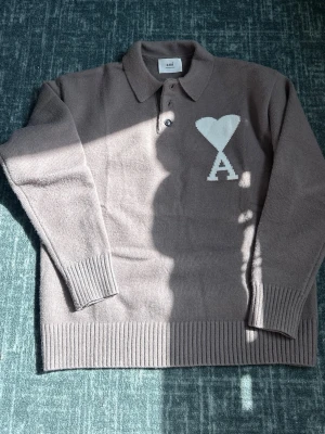 Stickad tröja med krage från Ami - Säljer en stickad tröja från Ami Alexandre Mattiussi i en mjuk, beige nyans. Tröjan har krage, knäppning framtill och ett stort vitt hjärta med bokstaven A på bröstet. Ribbstickade muddar och nederkant ger en snygg passform. Passar M/L