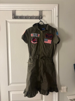 Top Gun klänning - Cool grön pilotdräkt inspirerad av Top Gun med flera broderade patchar, bland annat amerikansk flagga och Top Gun-logga. 