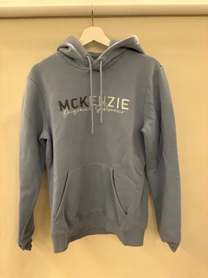 Ljusblå hoodie från McKenzie - Ljusblå hoodie från McKenzie med tryckt logga i vitt och brunt på bröstet. Klassisk känguruficka framtill, ribbades muddar och justerbar dragsko i huvan. Mjuk bomullsblandning som känns skön mot huden. Perfekt för en avslappnad streetstyle. 