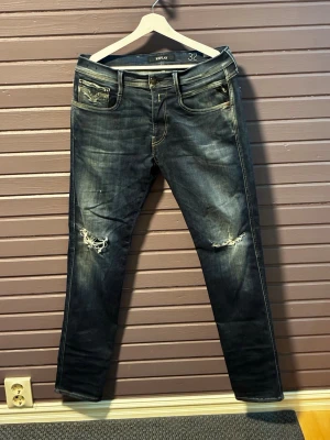 Replay Anbass slitna mörkblå jeans herr - Säljer ett par mörkblå Anbass jeans från Replay med slitningar och hål på båda knäna. Jeansen har klassisk femficksmodell, raka ben och tydliga tvättade detaljer för en cool, avslappnad look. Perfekta för dig som gillar grishstilen.