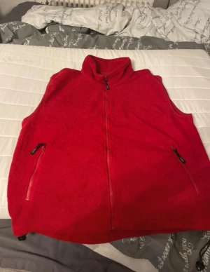 Röd fleeceväst med dragkedja - Riktigt skön röd fleeceväst med hel dragkedja framtill och två fickor med dragkedja. Hög krage för extra värme och enkel, sportig look. Perfekt att slänga på sig över en hoodie eller tröja.