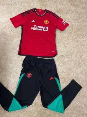 Manchester United träningsset Adidas - Snyggt träningsset från Adidas med Manchester Uniteds logga. Tröjan är röd med svarta detaljer och TeamViewer-tryck, byxorna är svarta med gröna paneler och röda loggor. Perfekt för fotbollsträning eller gymmet.