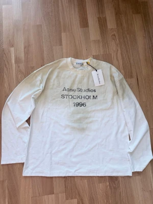 Vit långärmad topp Acne Studios - Vit långärmad topp från Acne Studios med tryck 'STOCKHOLM 1996' framtill. Klassisk rund halsringning och avslappnad passform. Perfekt för en clean och stilren look.