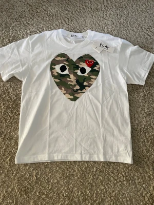  Comme des Garçons Play t-shirt - Vit t-shirt från Comme des Garçons Play med stort hjärta i camouflage-mönster och broderat rött hjärta med ögon på bröstet. Storlek M men passar som S