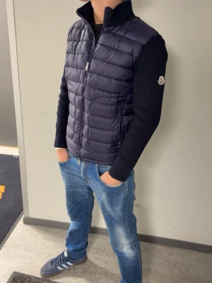 Mörkblå dunväst från Moncler - Snygg mörkblå dunväst från Moncler med quiltad front och rygg, samt stickade ärmar. Västen har dragkedja framtill, två dragkedjefickor och en hög krage. Klassisk Moncler-logga på ärmen och serietidningsdetalj på insidan.