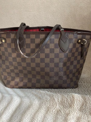 Louis Vuitton Neverfull Damier Ebene - Snygg handväska från Louis Vuitton i klassiskt Damier Ebene-mönster. Väskan är brun med rutigt mönster, har mörkbruna läderhandtag och detaljer i guld. Insidan är röd och väskan har en rymlig tote-form som passar till mycket.