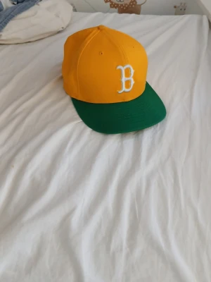 Gul och grön keps New Era 9FIFTY - Snygg snapback-keps från New Era 9FIFTY med gul krona, grön skärm och vit broderad B på framsidan. Klassisk basebollstil och justerbar passform. Perfekt accessoar för en sportig look.